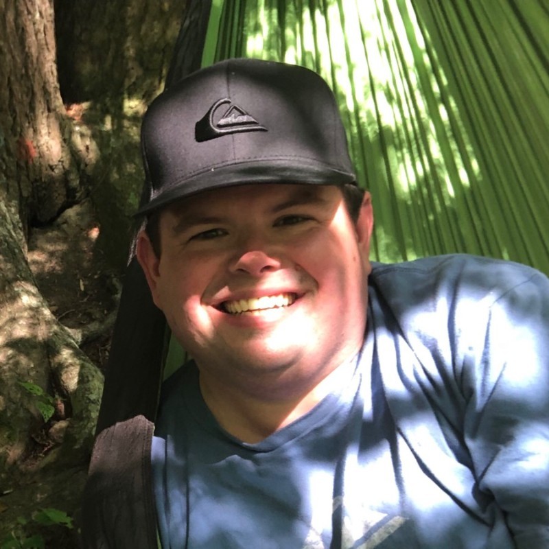 Steven wearing a black cap and a blue shirt, sitting in a green hammock surrounded by trees.